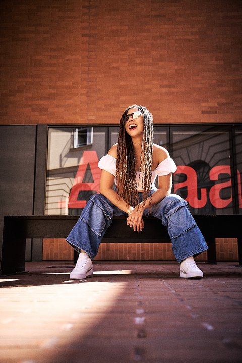 A woman with long, braided hair sits on a black bench, wearing a white off-the-shoulder top and wide-legged jeans. She smiles joyfully, surrounded by a sunlit outdoor setting, with a brick wall in the background featuring a large red sign.