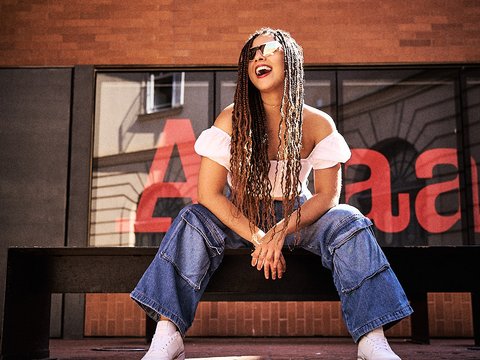 Eine junge Frau mit langen Dreadlocks sitzt lächelnd auf einer Bank. Sie trägt ein offenes, weißes Oberteil und weite, blaue Jeans. Die Sonne scheint auf sie, während sie entspannt und glücklich wirkt. Im Hintergrund sind Wände aus rotem Ziegel sichtbar.
