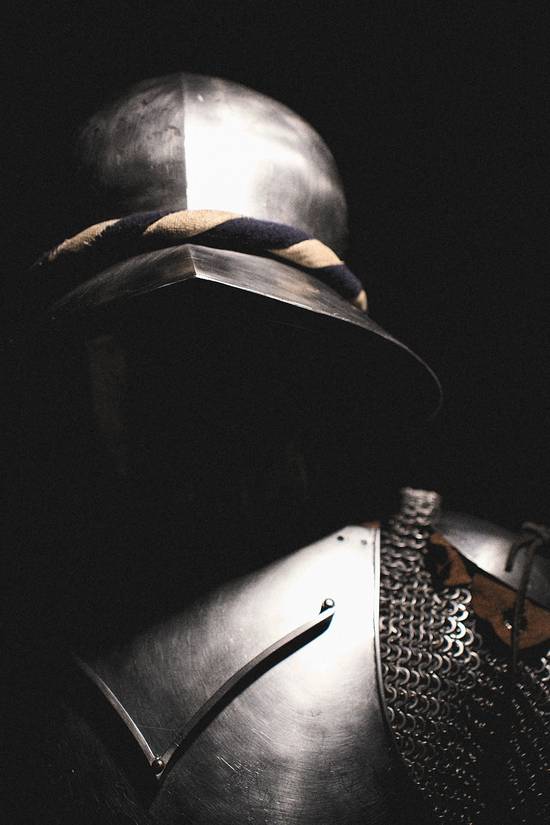 Ein metallener Ritterhelm mit einer charakteristischen Verzierung ist im Vordergrund sichtbar. Der Rest der Rüstung ist teilweise im Schatten, was eine mysteriöse Atmosphäre schafft. Die dunkle Umgebung hebt die glänzende Oberfläche des Helms hervor.