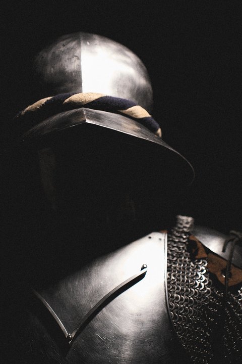 Ein metallener Ritterhelm mit einer charakteristischen Verzierung ist im Vordergrund sichtbar. Der Rest der Rüstung ist teilweise im Schatten, was eine mysteriöse Atmosphäre schafft. Die dunkle Umgebung hebt die glänzende Oberfläche des Helms hervor.