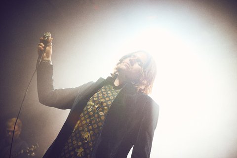 Ein Sänger steht auf der Bühne und hält ein Mikrofon in der Hand. Er trägt ein dunkelblaues Sakko und ein gemustertes Hemd. Im Hintergrund ist schwaches, diffuses Licht zu sehen. Die Szene vermittelt eine musikalische oder künstlerische Performance.