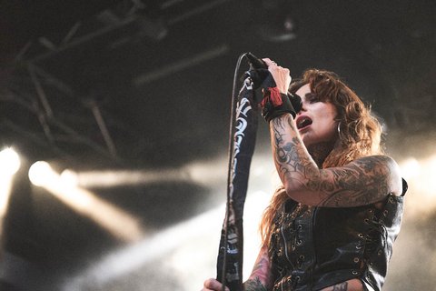 Eine tätowierte Sängerin steht auf der Bühne und singt in ein Mikrofon. Sie trägt ein schwarzes, ärmelloses Oberteil und hat lange lockige Haare. Im Hintergrund sind Scheinwerfer, die ein schwaches Licht ausstrahlen. Die Atmosphäre ist energisch und musikalisch.