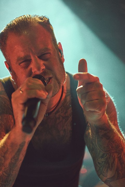 Ein männlicher Sänger mit tätowierten Armen performt auf der Bühne. Er hält ein Mikrofon und zeigt mit dem Daumen nach oben. Die Beleuchtung ist diffus und schafft eine energiegeladene Atmosphäre.