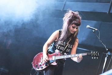 Eine Musikerin mit langen, dichten Haaren spielt begeistert eine rote E-Gitarre. Sie trägt ein schwarzes T-Shirt mit einem feministischen Slogan und steht auf einer Bühne, umgeben von Nebel und Licht. Ihre Mimik zeigt Konzentration und Freude am Musizieren.