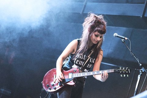 Eine Musikerin mit langen, dichten Haaren spielt begeistert eine rote E-Gitarre. Sie trägt ein schwarzes T-Shirt mit einem feministischen Slogan und steht auf einer Bühne, umgeben von Nebel und Licht. Ihre Mimik zeigt Konzentration und Freude am Musizieren.