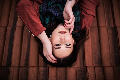 Overhead shot of a young woman with dark hair, striking blue eyes, and dramatic winged eyeliner, lying on her back on terracotta tiles. She wears a rust-colored jacket and plaid scarf, her hands gently framing her face, looking directly at the viewer.