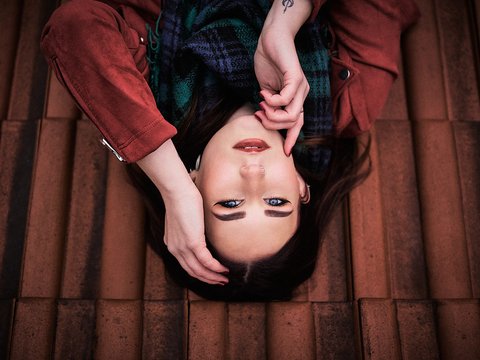 Eine junge Frau liegt auf einer Fliese, sieht zur Kamera und berührt ihr Gesicht mit einer Hand. Sie trägt einen Schal und hat langes, dunkles Haar. Der Hintergrund besteht aus roten Fliesen. Der Ausdruck ihres Gesichts ist nachdenklich.