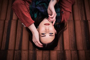 Eine junge Frau liegt auf einer Fliese, sieht zur Kamera und berührt ihr Gesicht mit einer Hand. Sie trägt einen Schal und hat langes, dunkles Haar. Der Hintergrund besteht aus roten Fliesen. Der Ausdruck ihres Gesichts ist nachdenklich.