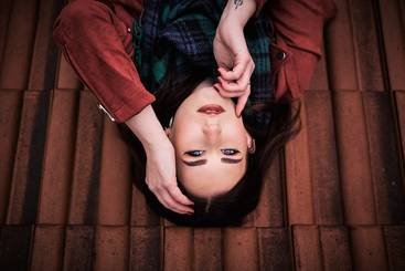 Eine junge Frau liegt auf einer Fliese, sieht zur Kamera und berührt ihr Gesicht mit einer Hand. Sie trägt einen Schal und hat langes, dunkles Haar. Der Hintergrund besteht aus roten Fliesen. Der Ausdruck ihres Gesichts ist nachdenklich.