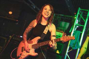 Eine lächelnde Musikerin spielt Gitarre auf einer Bühne. Sie trägt ein schwarzes Outfit und hat einen sichtbaren Tattoos-Ärmel. Die Beleuchtung ist farbenfroh, was die lebendige Atmosphäre des Auftritts unterstreicht.