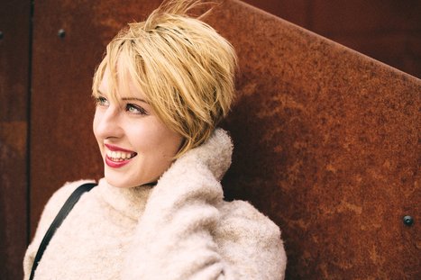 A smiling woman with short, tousled blonde hair is leaning against a textured background. She wears a light-colored, cozy sweater and appears relaxed and happy. Her expression is cheerful, with bright eyes and a warm smile.