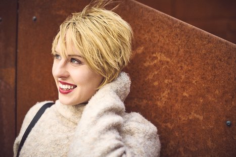 Eine lächelnde Frau mit kurzen, blonden Haaren trägt einen hellen Pullover. Sie lehnt entspannt gegen eine rostige Wand und hat eine Hand an ihrem Hals. Ihr Gesicht strahlt Freude und Zufriedenheit aus.