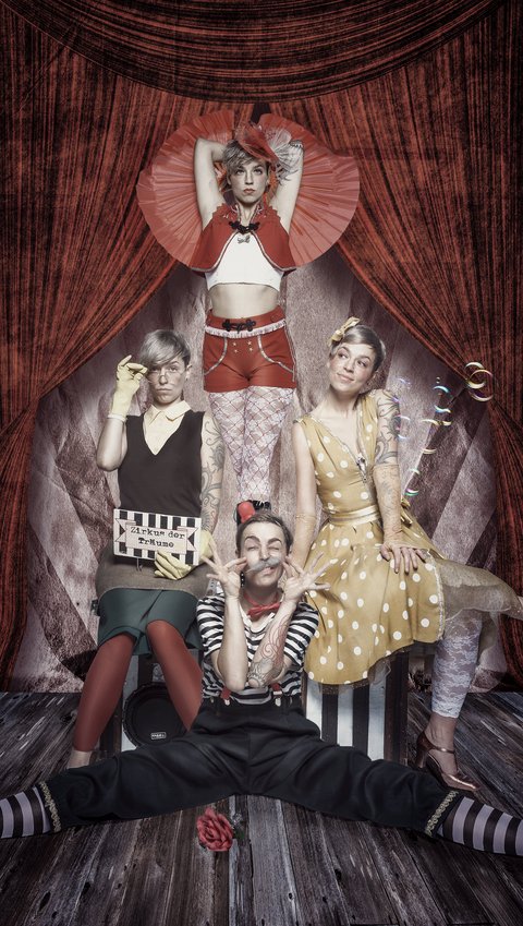 Vier Menschen in bunten Kostümen posieren kreativ vor einem roten Vorhang. Ein Clown sitzt auf dem Boden, während die anderen in unterschiedlichen Posen und Outfits stehen und sitzen, mit einem Zugang zur Theateratmosphäre. Die Szene vermittelt Freude und Mysterium.