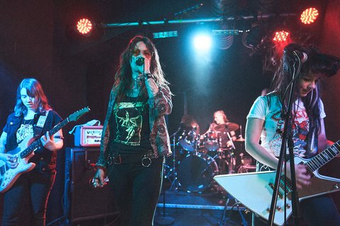 Eine Rockband tritt auf einer Bühne auf. Die Sängerin mit Mikrofon in der Mitte wird von zwei Gitarristinnen flankiert, während ein Schlagzeuger im Hintergrund spielt. Die Atmosphäre ist energiegeladen und die Band trägt rocktypische Kleidung. Rote und blaue Lichter beleuchten die Szenerie.