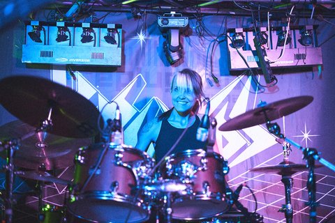 Ein Schlagzeuger spielt leidenschaftlich auf der Bühne. Er lächelt und ist von bunten Lichtern umgeben, die die energische Atmosphäre des Auftritts unterstreichen. Im Hintergrund sind Drum-Sets und Scheinwerfer sichtbar, was die musikalische Darbietung hervorhebt.
