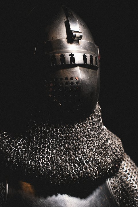 Ein mittelalterlicher Ritter in glänzender Rüstung und einem geschlossenen Helm. Die Rüstung ist aus Kettenhemd gefertigt, das den Halsbereich schützt. Das Bild zeigt den Ritter halbseitig, mit einem dunklen Hintergrund, der die Rüstung hervorhebt.