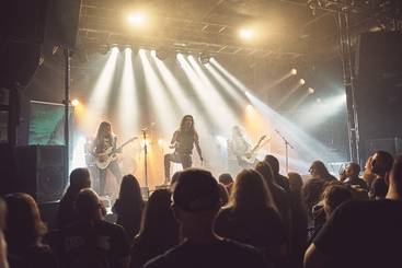 Ein Live-Konzert mit einer Band auf der Bühne. Der Sänger steht im Vordergrund, während die Musiker im Hintergrund spielen. Hell erleuchtete Scheinwerfer werfen Lichtstrahlen auf die Bühne. Die Menge im Publikum schaut gebannt zu.