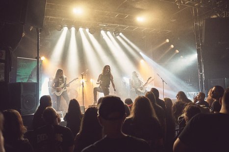 A live music performance takes place on stage, featuring a band with a lead vocalist and musicians playing guitars. Bright stage lights illuminate the scene, while an enthusiastic crowd watches closely, creating an energetic atmosphere.