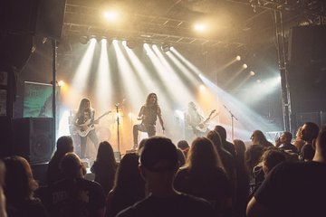 Ein Live-Konzert mit einer Band auf der Bühne. Der Sänger steht im Vordergrund, während die Musiker im Hintergrund spielen. Hell erleuchtete Scheinwerfer werfen Lichtstrahlen auf die Bühne. Die Menge im Publikum schaut gebannt zu.