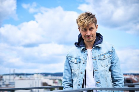 Ein Mann mit blonden, leicht zerzausten Haaren steht an einem Geländer. Er trägt eine hellblaue Jeansjacke über einem weißen T-Shirt und schaut lächelnd in die Kamera. Im Hintergrund sind himmelblaue Wolken und eine Stadtlandschaft zu sehen.