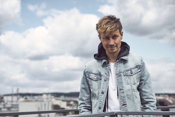 Ein Mann mit blonden, leicht zerzausten Haaren steht an einem Geländer. Er trägt eine hellblaue Jeansjacke über einem weißen T-Shirt und schaut lächelnd in die Kamera. Im Hintergrund sind himmelblaue Wolken und eine Stadtlandschaft zu sehen.
