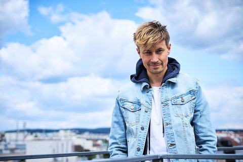 A young man with tousled blond hair stands on a balcony, wearing a light denim jacket over a white shirt. He gazes slightly to the side, with a soft smile, against a backdrop of a blue sky and scattered clouds. The city skyline is visible in the distance.