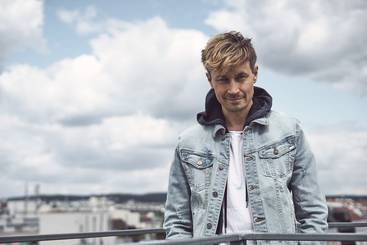 Ein Mann mit blonden, leicht zerzausten Haaren steht an einem Geländer. Er trägt eine hellblaue Jeansjacke über einem weißen T-Shirt und schaut lächelnd in die Kamera. Im Hintergrund sind himmelblaue Wolken und eine Stadtlandschaft zu sehen.