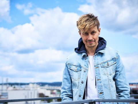 Ein Mann mit blonden, leicht zerzausten Haaren steht an einem Geländer. Er trägt eine hellblaue Jeansjacke über einem weißen T-Shirt und schaut lächelnd in die Kamera. Im Hintergrund sind himmelblaue Wolken und eine Stadtlandschaft zu sehen.