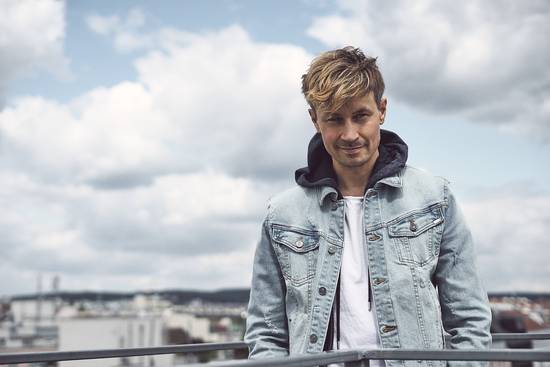 Ein Mann mit blonden, leicht zerzausten Haaren steht an einem Geländer. Er trägt eine hellblaue Jeansjacke über einem weißen T-Shirt und schaut lächelnd in die Kamera. Im Hintergrund sind himmelblaue Wolken und eine Stadtlandschaft zu sehen.