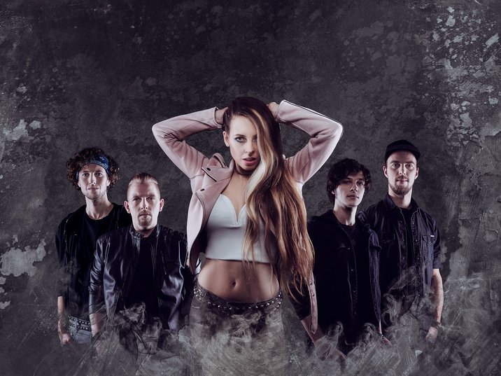 A music band is posing in front of a textured backdrop. The lead singer, a woman with long hair, stands confidently in front, with her hands behind her head. Four male band members are positioned behind her, all dressed in dark attire.