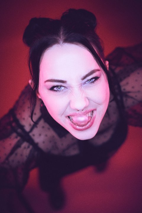 A person with dark hair styled in two buns is seen from above, expressing excitement or joy. They wear a sheer black top with polka dots and are surrounded by a vibrant pink background, enhancing their energetic expression.