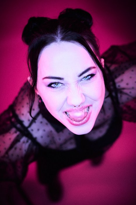 Fierce Gaze Under Magenta Light A person with dark hair styled in two buns is seen from above, expressing excitement or joy. They wear a sheer black top with polka dots and are surrounded by a vibrant pink background, enhancing their energetic expression.