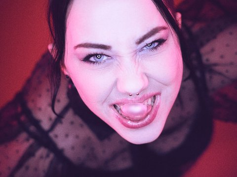 Eine Person mit dunklem, transparentem Oberteil zeigt ein breites Lächeln und spricht energetisch in die Kamera. Ihr Gesicht ist von einem pinken Hintergrund beleuchtet. Die Stimmung wirkt lebhaft und ausdrucksstark.