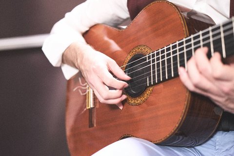 Ein Musiker spielt auf einer akustischen Gitarre. Die Hände sind auf den Saiten positioniert, während er eine Melodie spielt. Der Fokus liegt auf der handwerklichen Bewegung und der Konstruktion der Gitarre.
