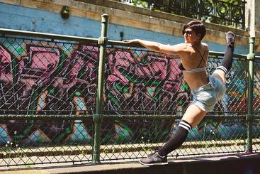 Eine sportliche Person dehnt sich an einem Metallzaun, umgeben von buntem Graffiti. Sie trägt ein Sportoutfit mit kurzer Hose und Socken über dem Knie, während sie mit einer Hand am Zaun stützt. Die Pose vermittelt Flexibilität und Energie.