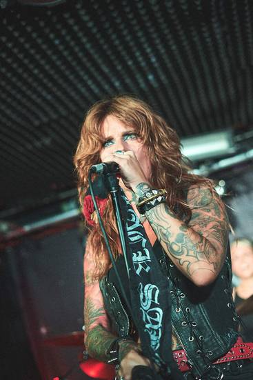 Eine Sängerin mit langen, gewellten, braunen Haaren steht auf der Bühne und singt ins Mikrofon. Sie trägt eine schwarze Lederjacke, hat zahlreiche Tattoos und sieht leidenschaftlich aus. Im Hintergrund sind verschwommen weitere Musiker zu erkennen, die an Instrumenten spielen.