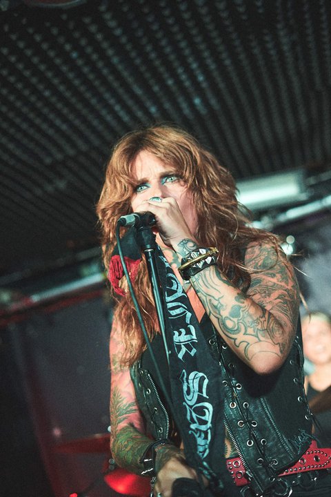 Eine Sängerin mit langen, gewellten, braunen Haaren steht auf der Bühne und singt ins Mikrofon. Sie trägt eine schwarze Lederjacke, hat zahlreiche Tattoos und sieht leidenschaftlich aus. Im Hintergrund sind verschwommen weitere Musiker zu erkennen, die an Instrumenten spielen.