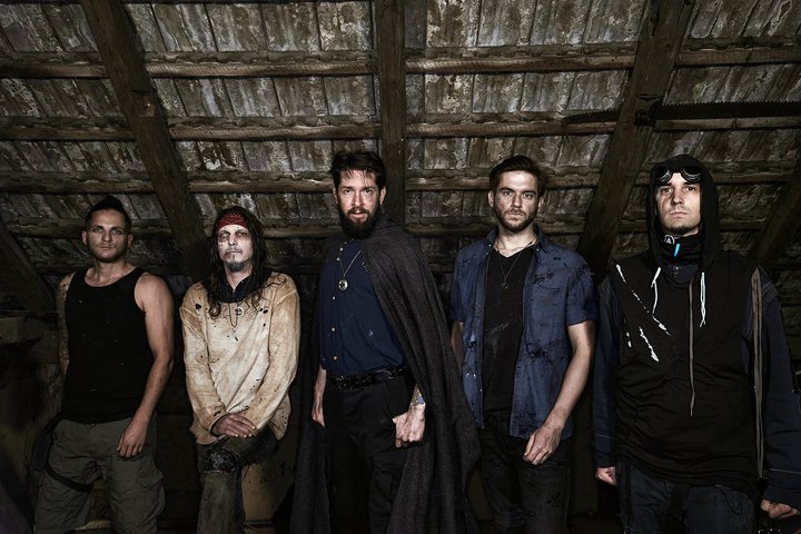 Fünf Männer stehen in einem düsteren Raum mit Holzbalkendecke. Jeder trägt unterschiedliche Freizeitkleidung, einige haben auffällige Gesichtsbemalung oder Kleidung. Die Stimmung ist geheimnisvoll und etwas unheimlich, was durch die dunklen Farben und die Lichtverhältnisse verstärkt wird.