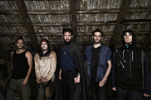 A group of five individuals stands together in a dimly lit attic. They are dressed in various dark, unconventional outfits, exuding a mysterious vibe. The atmosphere suggests a cohesive theme, possibly related to music or performance. The background features wooden beams and a rustic ceiling.
