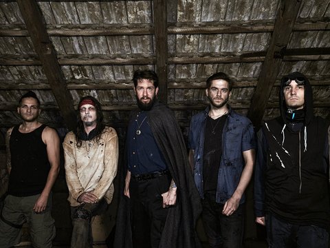 Fünf Männer stehen in einem düsteren Raum mit Holzbalkendecke. Jeder trägt unterschiedliche Freizeitkleidung, einige haben auffällige Gesichtsbemalung oder Kleidung. Die Stimmung ist geheimnisvoll und etwas unheimlich, was durch die dunklen Farben und die Lichtverhältnisse verstärkt wird.