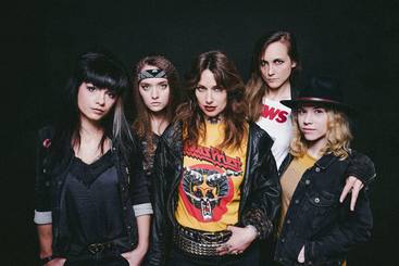 Fünf Frauen stehen zusammen in einer Gruppe, gekleidet in modische, rockige Outfits. Sie zeigen individuelle Stile mit Lederjacken, T-Shirts und Accessoires, während sie intensiv in die Kamera blicken. Der Hintergrund ist dunkel, was den Fokus auf die Gruppe verstärkt.