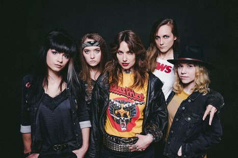 Fünf Frauen stehen zusammen in einer Gruppe, gekleidet in modische, rockige Outfits. Sie zeigen individuelle Stile mit Lederjacken, T-Shirts und Accessoires, während sie intensiv in die Kamera blicken. Der Hintergrund ist dunkel, was den Fokus auf die Gruppe verstärkt.