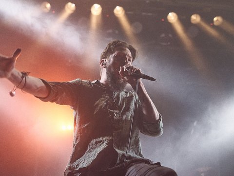 Ein Sänger steht auf der Bühne und singt in ein Mikrofon. Er hat die Arme weit ausgestreckt und wirkt leidenschaftlich. Die Bühne ist mit buntem Licht beleuchtet, was eine energiegeladene Atmosphäre schafft.