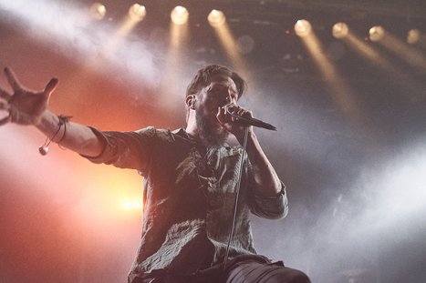 A male singer passionately performs on stage, holding a microphone and gesturing energetically. Bright stage lights create a dramatic atmosphere, highlighting his expressive movements as he engages with the audience.