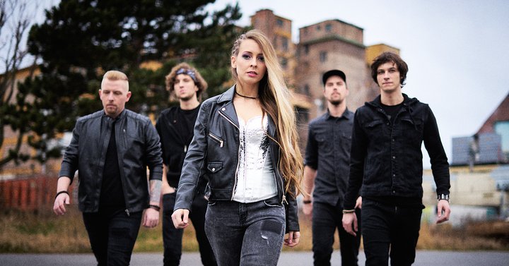 Eine fünfköpfige Band steht in aufrechter Haltung auf einem Weg. Die Sängerin in der Mitte trägt eine Lederjacke und hat lange Haare. Die anderen Bandmitglieder sind maskulin gekleidet und schauen ernst. Im Hintergrund sind verschwommene Gebäude zu sehen.