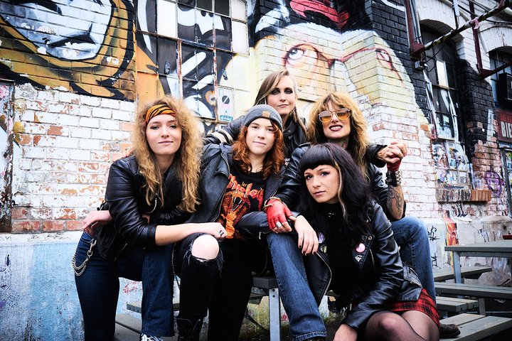 Fünf Frauen posieren lässig vor einer bemalten Wand. Sie tragen Lederjacken und haben unterschiedliche Frisuren, die ihren individuellen Stil unterstreichen. Die Atmosphäre ist selbstbewusst und freundlich.