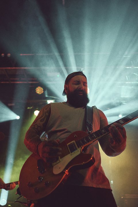 Ein Musiker mit Bart spielt E-Gitarre auf einer Bühne, umgeben von intensivem Licht. Die Strahlen von Scheinwerfern erzeugen eine dynamische Atmosphäre. Der Musiker trägt ein ärmelloses Oberteil und ist fokussiert auf sein Spiel.