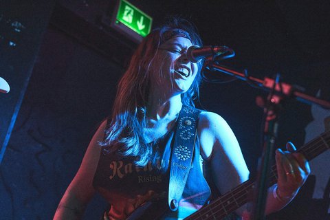 Eine Sängerin mit langen, offenem Haar steht am Mikrofon und spielt Bass. Sie wirkt energiegeladen und lächelt während des Auftritts. Der Raum ist schwach beleuchtet, wobei blaue Lichtakzente die Atmosphäre verstärken.