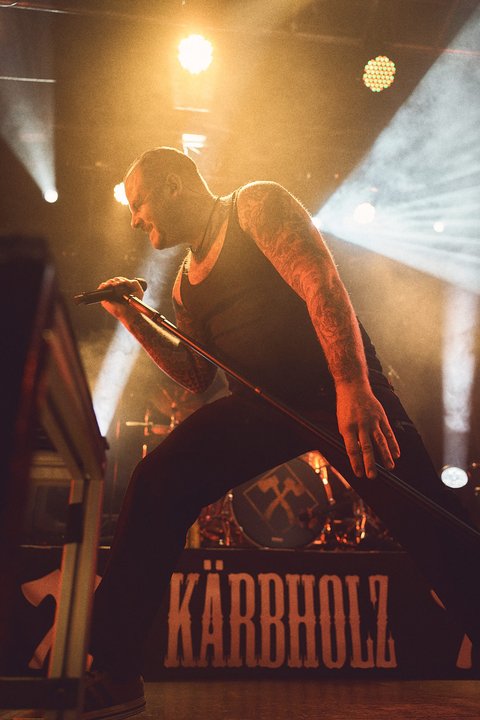 Ein Sänger steht auf einer Bühne und singt leidenschaftlich ins Mikrofon. Er trägt ein ärmelloses Shirt und hat tätowierte Arme. Im Hintergrund sind Lichtstrahlen zu sehen, die eine energiegeladene Atmosphäre erzeugen. Der Name der Band „Kärbholz“ ist deutlich sichtbar.