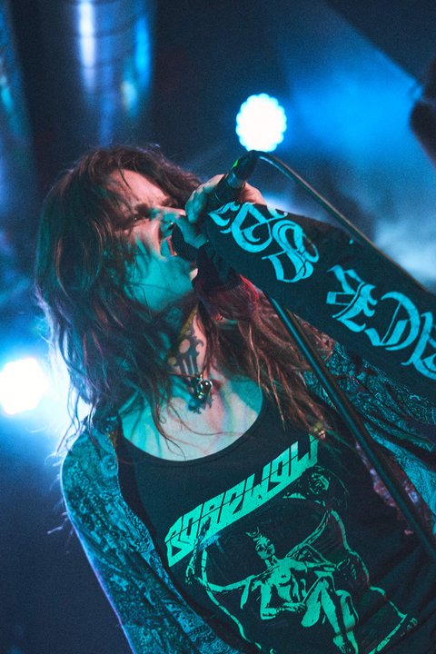 Eine Sängerin mit langen, lockigen Haaren steht auf der Bühne und singt in ein Mikrofon. Sie trägt ein schwarzes T-Shirt mit einem auffälligen Design und hat eine leidenschaftliche Ausstrahlung. Die Beleuchtung um sie herum schafft eine dynamische, energiegeladene Atmosphäre.